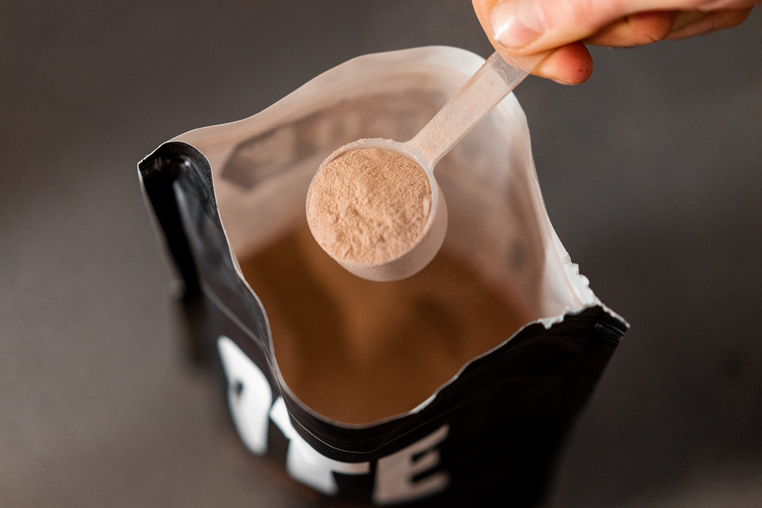 Person holding a scoop of protein powder above a bag labeled 'Protein'.