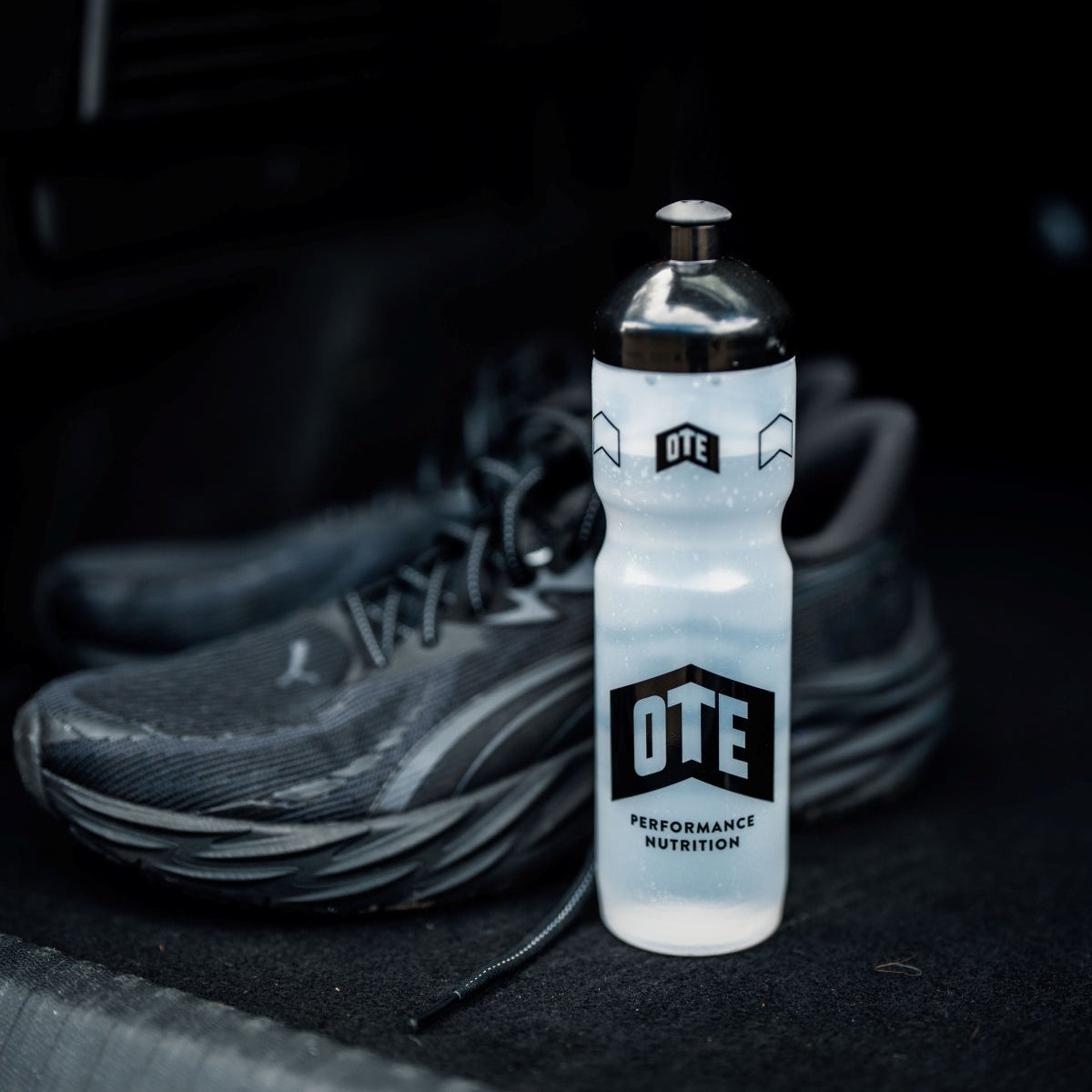 White water bottle with 'OTE Performance Nutrition' branding next to a black athletic shoe on a dark background