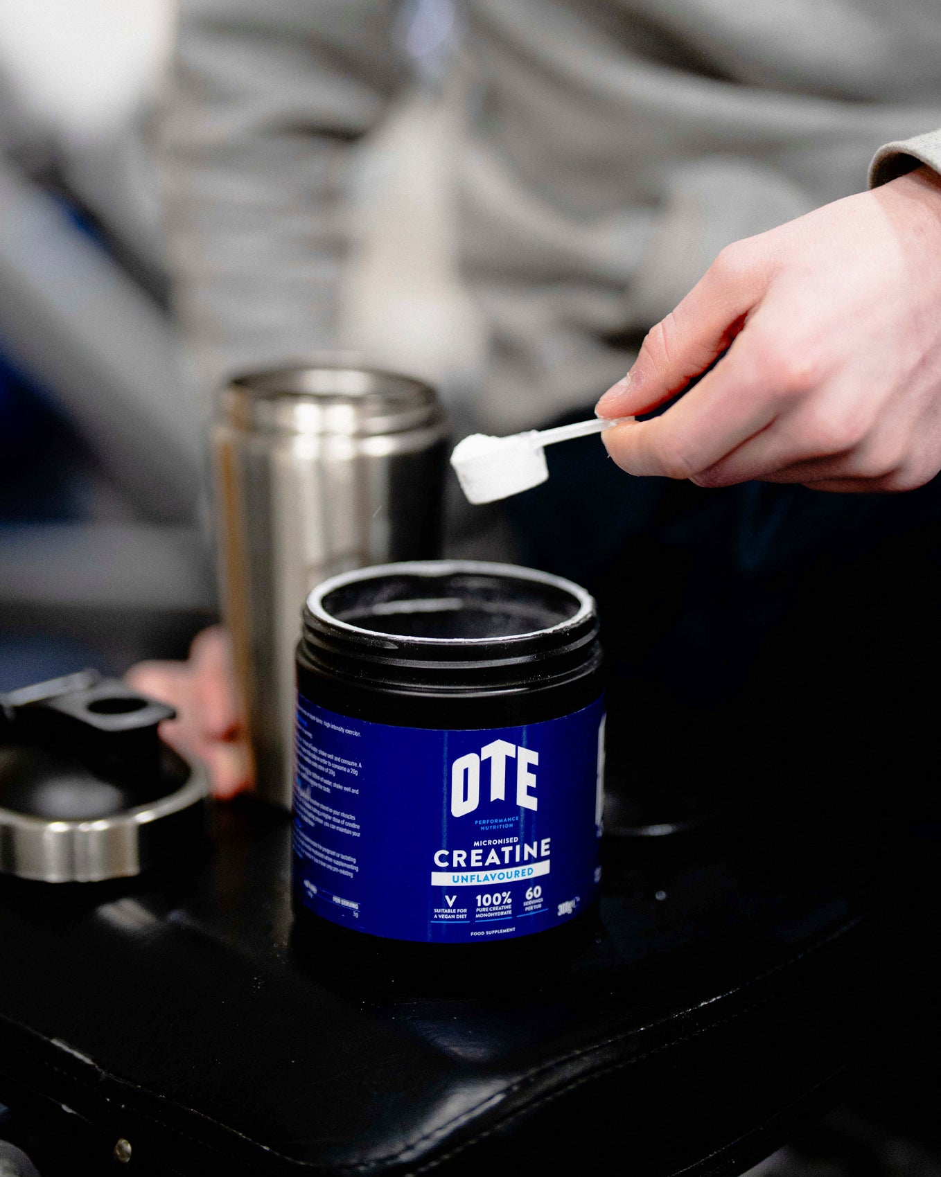 Person using a scoop to take creatine powder from a blue container labeled 'OTE Creatine' on a black surface.